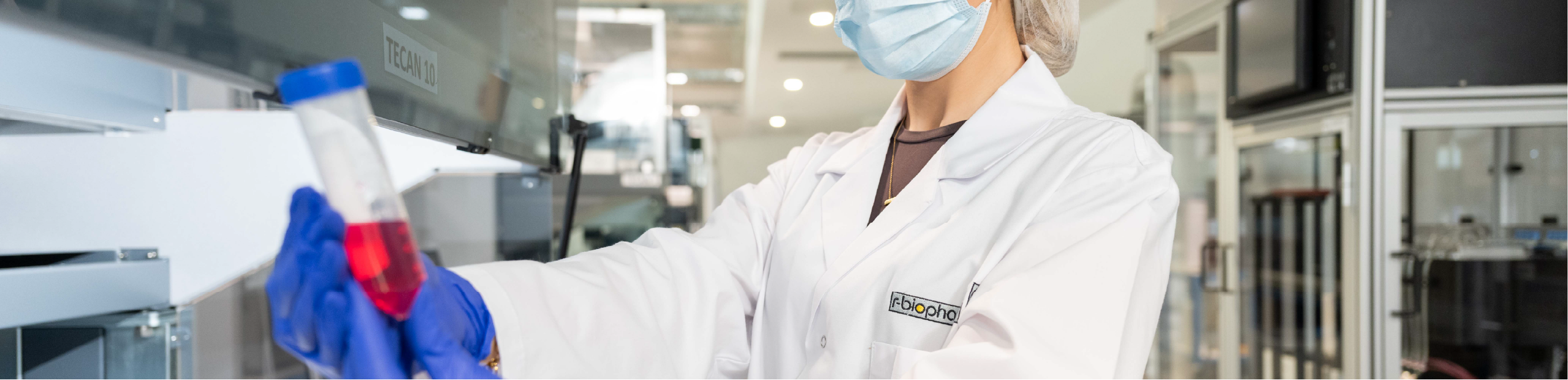 a woman wearing a hairnet, face mask, lab coat and gloves holding a tube of fluid for use in medical diagnostics