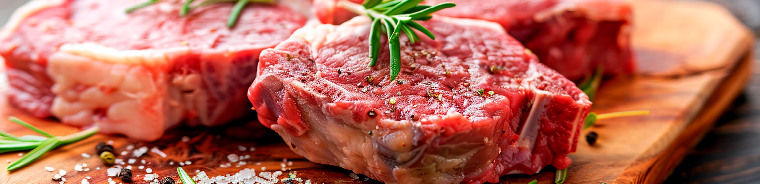 three raw steaks with fresh herbs, salt and pepper placed on top, sitting on a chopping board
