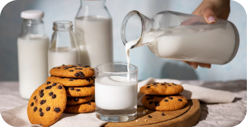 A person pours milk from a glass bottle into a small glass near chocolate chip cookies