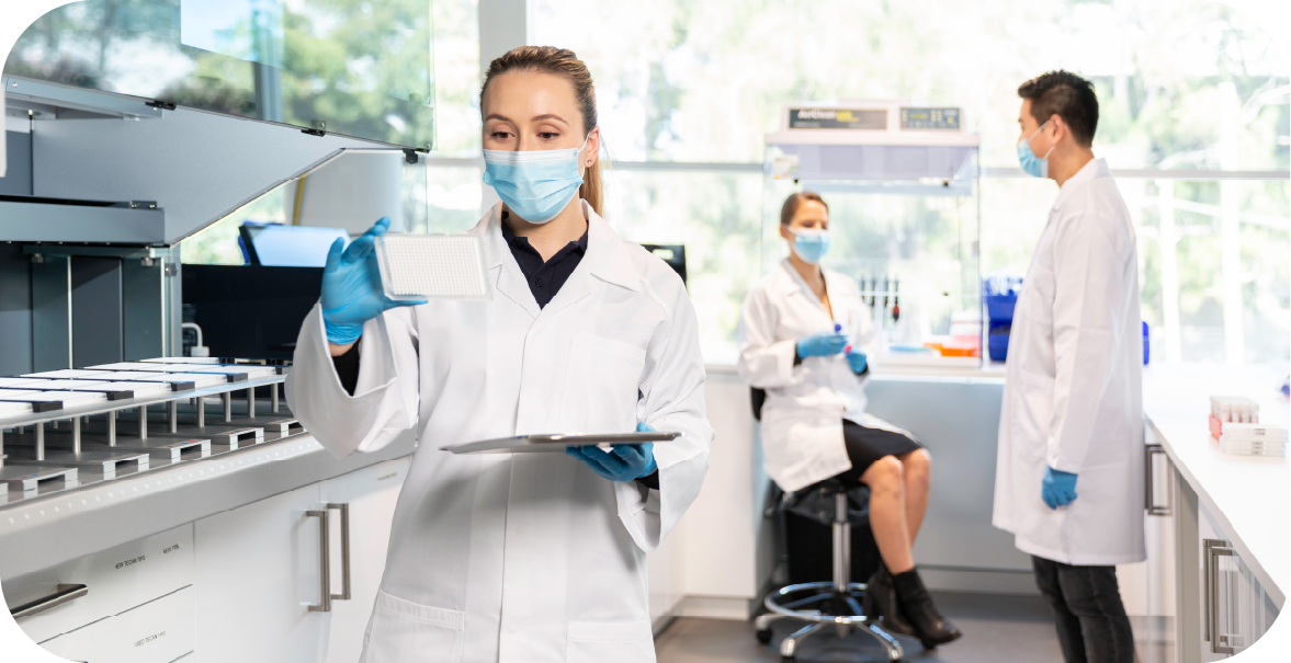 A woman in a lab coat holds a tray of respiratory pathogen tests while two scientists have an animated discussion in the background