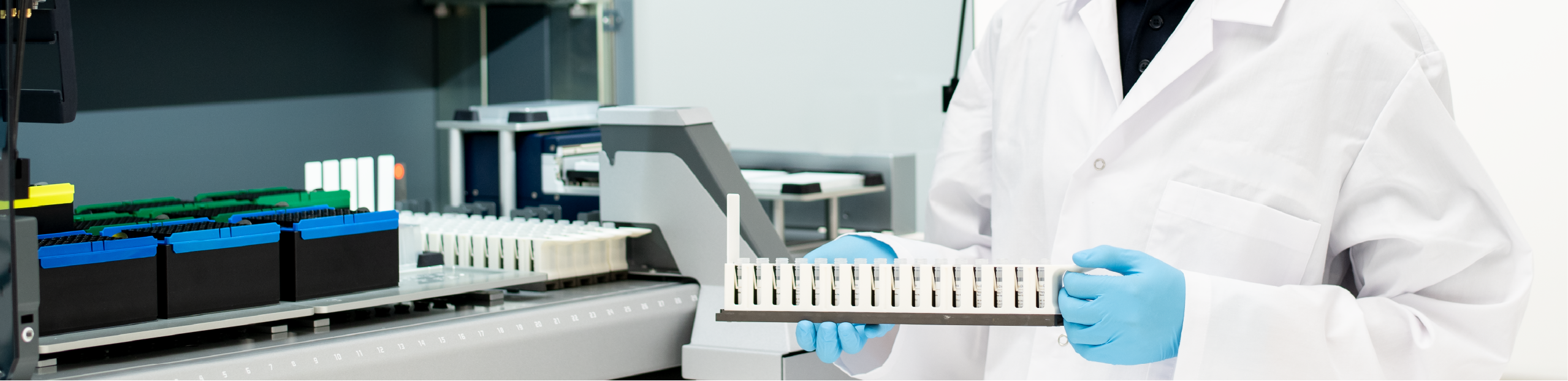 a man in a lab coat with gloves holding a tray of medical diagnostic tests in preparation to conduct diagnostic testing in a large instrument