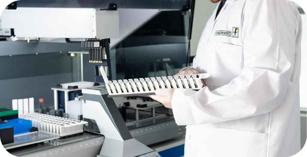 A man in a R-Biopharm branded lab coat holds a tray containing medical diagnostic equipment in front of a complex instrument designed to conduct the RIDA®Xtract 24 pathogen kit