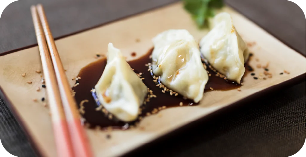 three dumplings sit in a pool of soy sauce on a rectangular plate with chopsticks