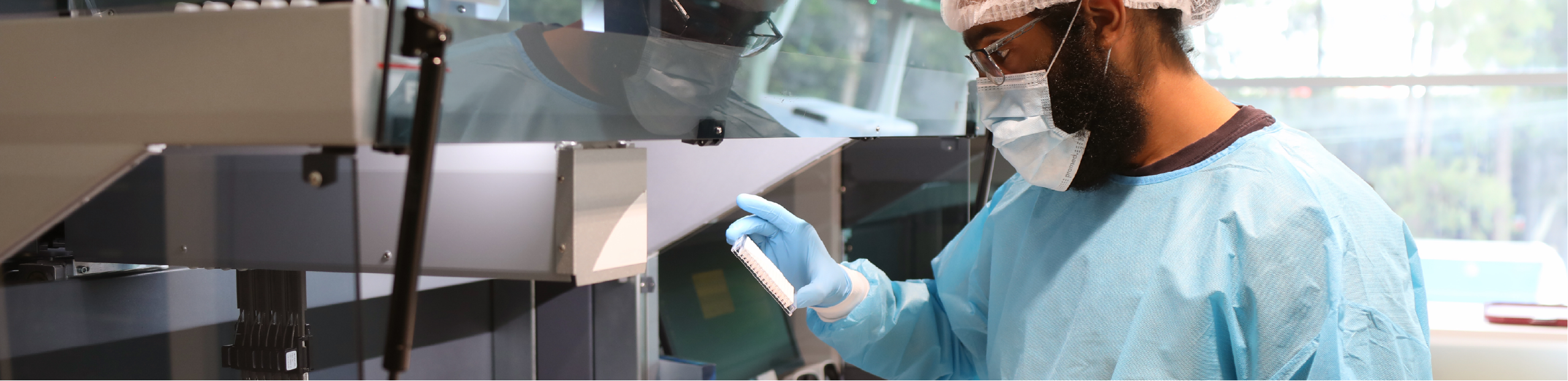 a man in a hair net, lab coat and gloves inspects a plate near a large piece of medical diagnostics equipment