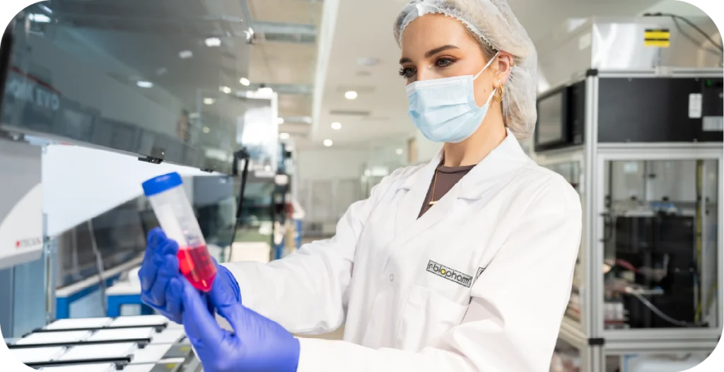a woman in a lab coat, hair net and gloves inspects a tube filled with red liquid in front of complex medical diagnostic machinery