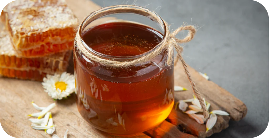 a jar of honey sits on a wooden chopping board with honeycomb behind it