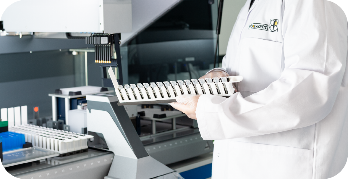 A man in a R-Biopharm branded lab coat holds a tray containing medical diagnostic equipment in front of a complex instrument designed to conduct medical diagnostic tests