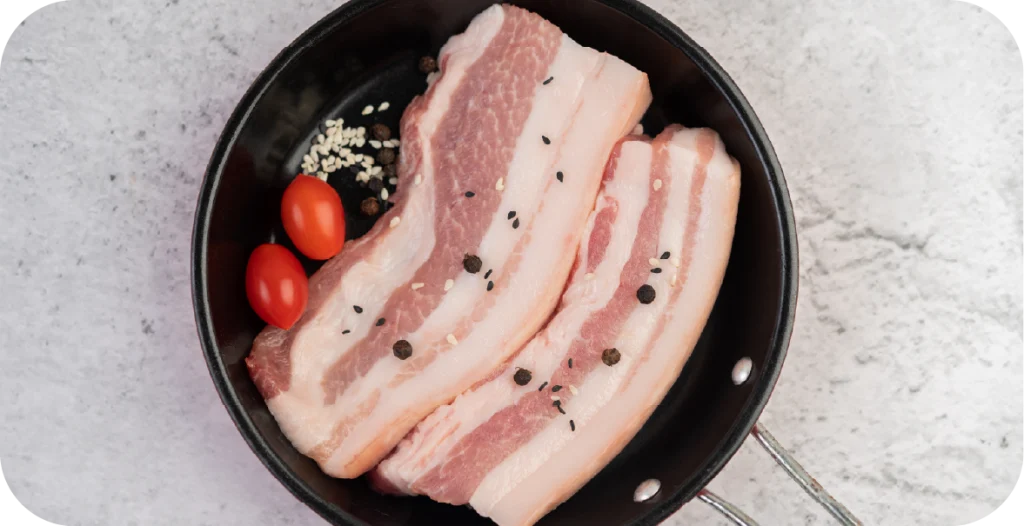 two thick strips of pork belly in a frying pan with peppercorns and cherry tomatoes