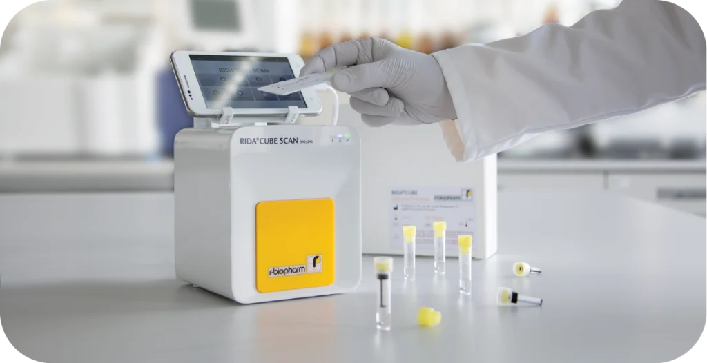 The RIDA®CUBE SCAN, a small white box with a yellow front and a slot for a device. A scientist places a lateral flow test near the reader