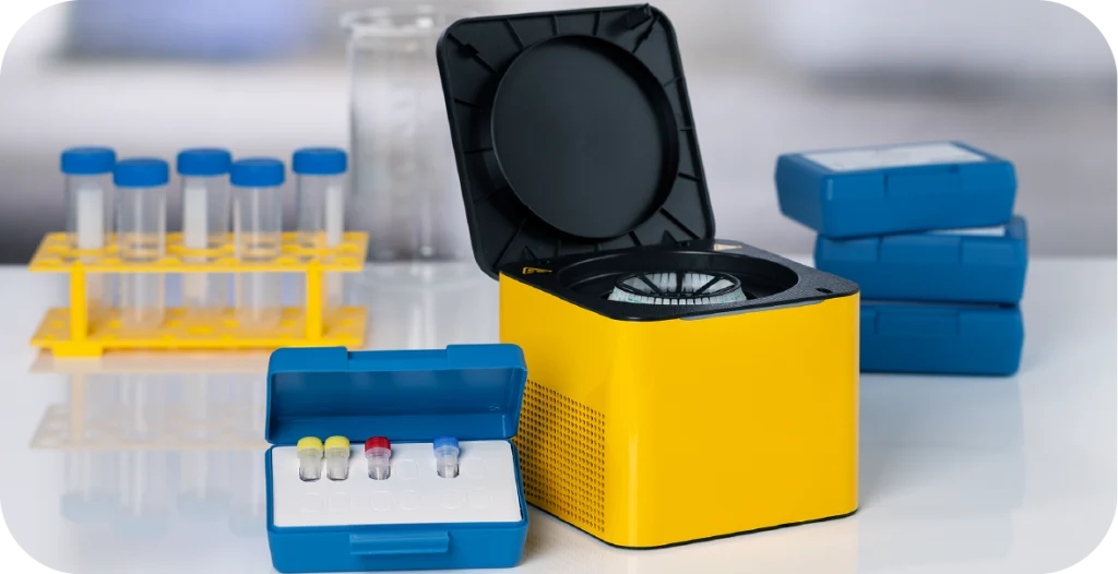 A small yellow box with a black lid sits behind a cassette tray and its lid, next to blue test packages and test tubes