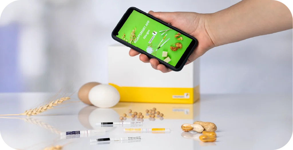 A person holding a phone with the RIDA®SMART APP Allergen in front of a diagnostic test and food items